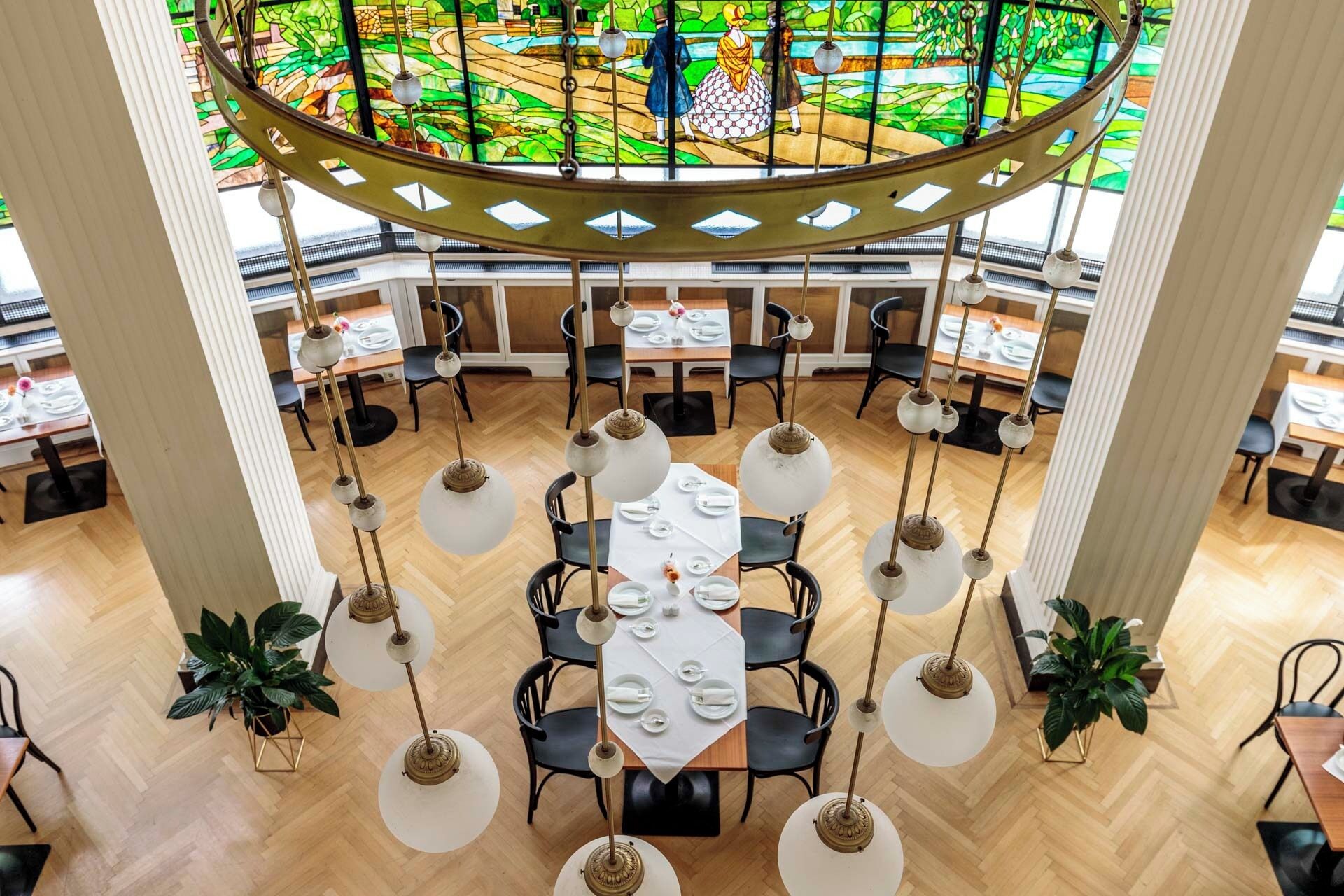 A spacious dining area with multiple tables and chairs, a central chandelier, and a stained glass window above.