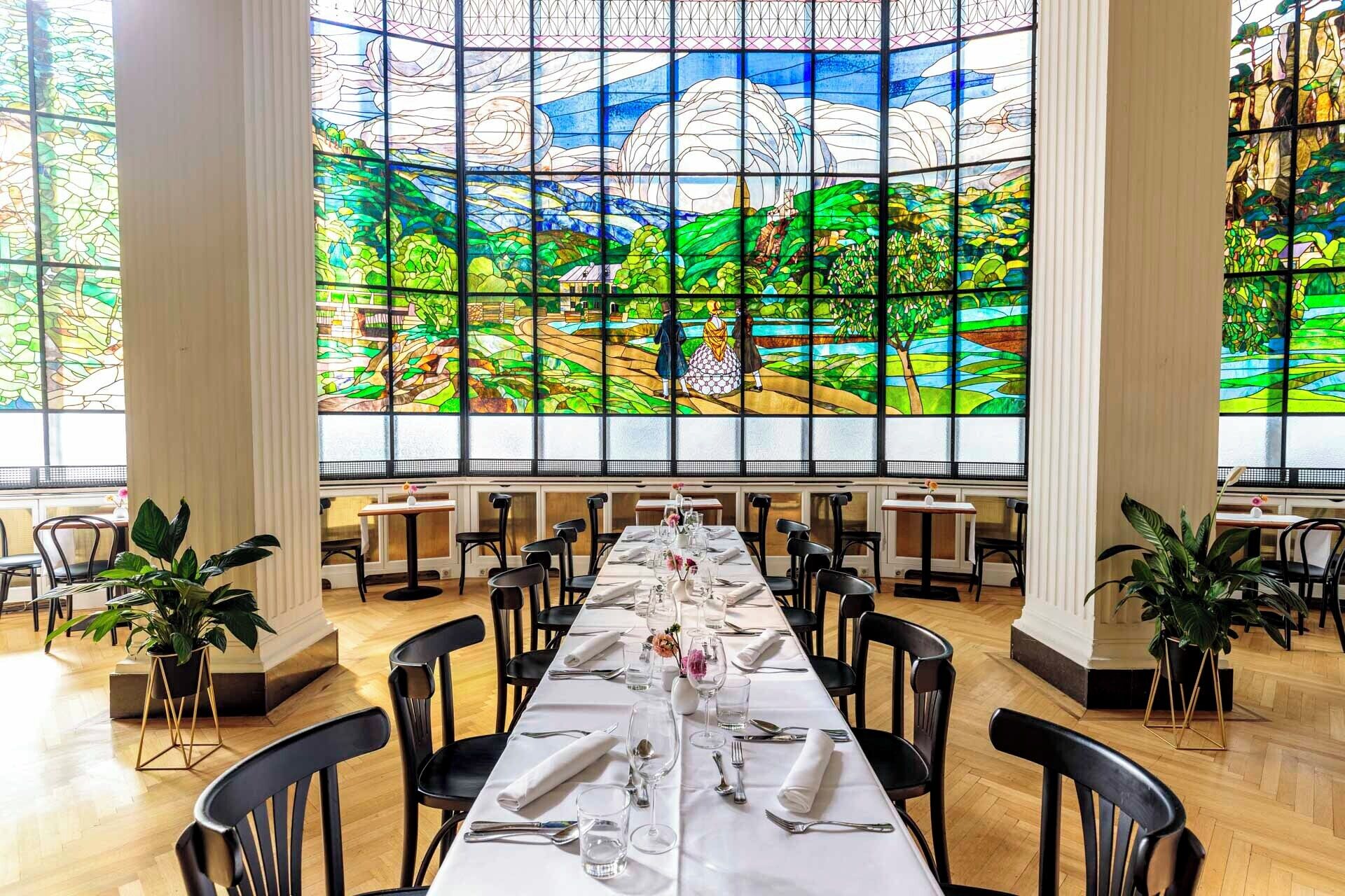 A dining area with a long table set for a meal, large stained glass windows, and potted plants.