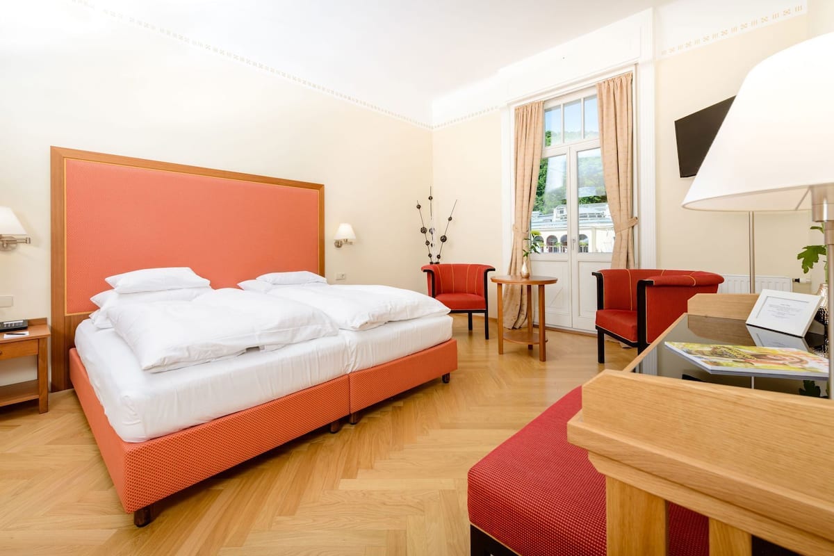 A hotel room with a large bed, two red chairs, a small table, and a desk with a lamp.
