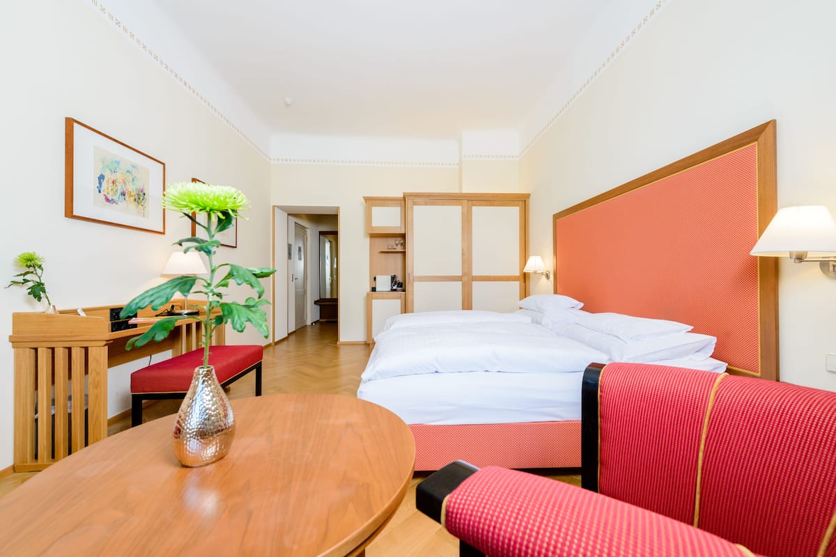 A hotel room with a bed, a desk, a chair, a table, and a potted plant.