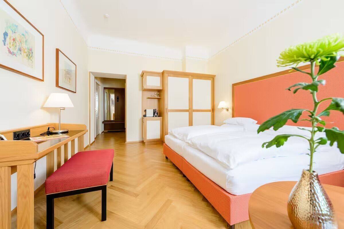 A hotel room with a bed, a desk with a chair, a bedside table, a lamp, and a plant.