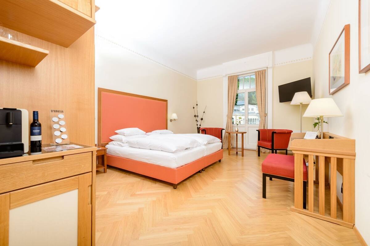 A hotel room with a bed, a desk with a coffee machine, a chair, a table with a vase, and a TV on the wall.