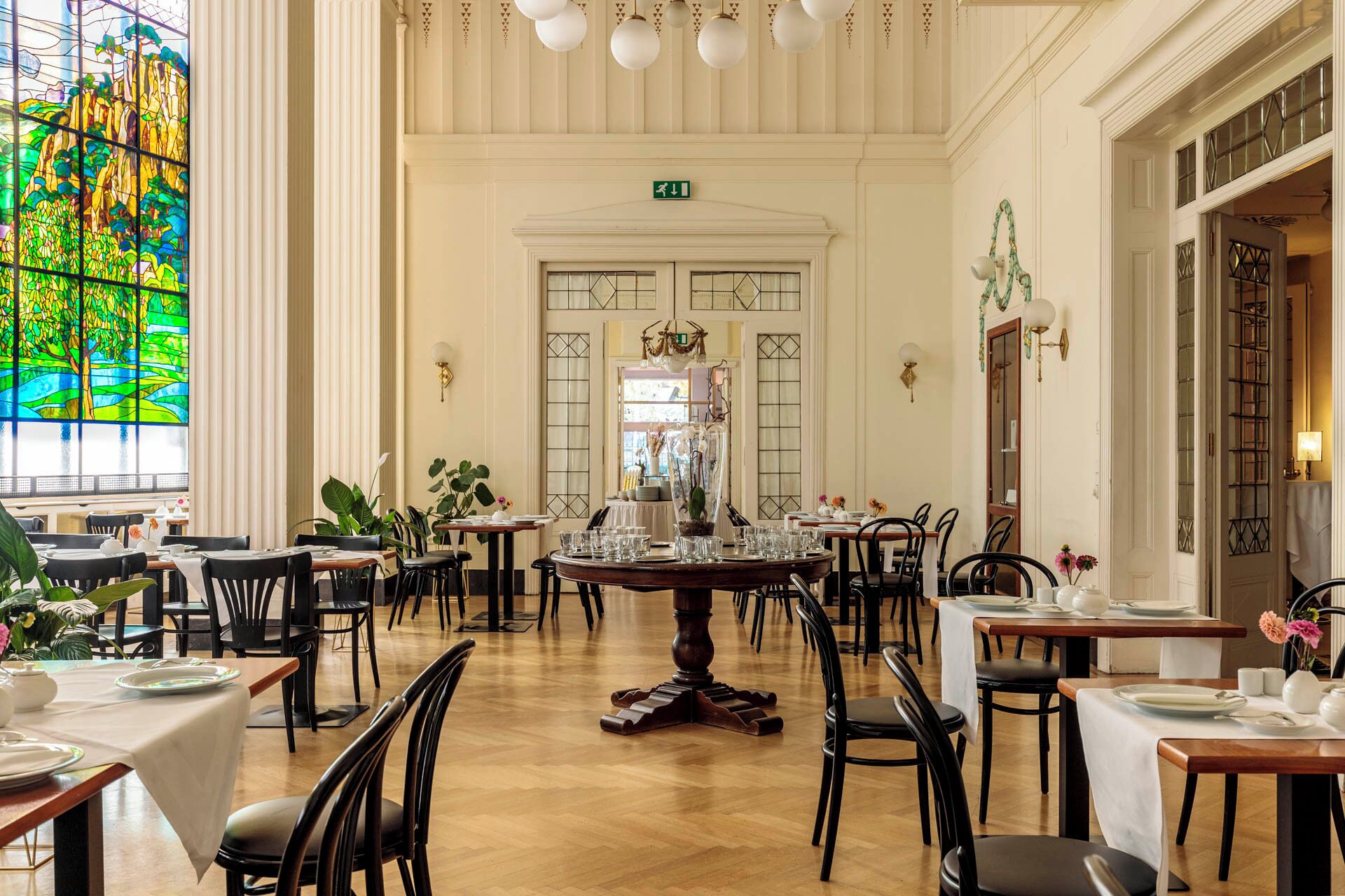 A spacious dining area with multiple tables and chairs, a large stained glass window, and a decorative chandelier.