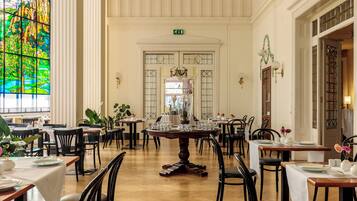 Un vaste espace dédié à la salle à manger, avec de nombreuses tables et chaises, une grande fenêtre à vitraux et un lustre décoratif.