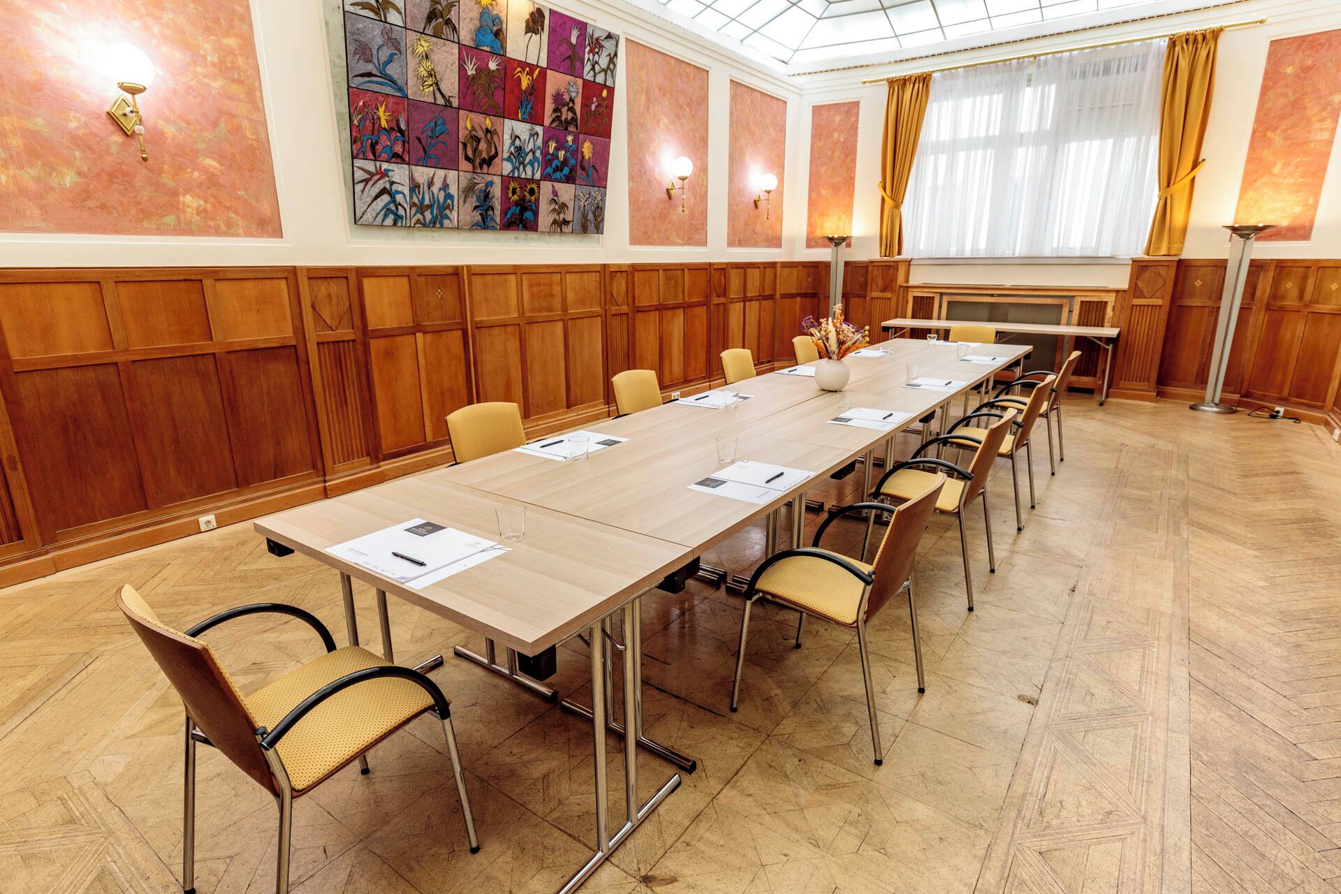 A conference room with a long table, chairs, wooden paneling, a large artwork on the wall, and a window with curtains.