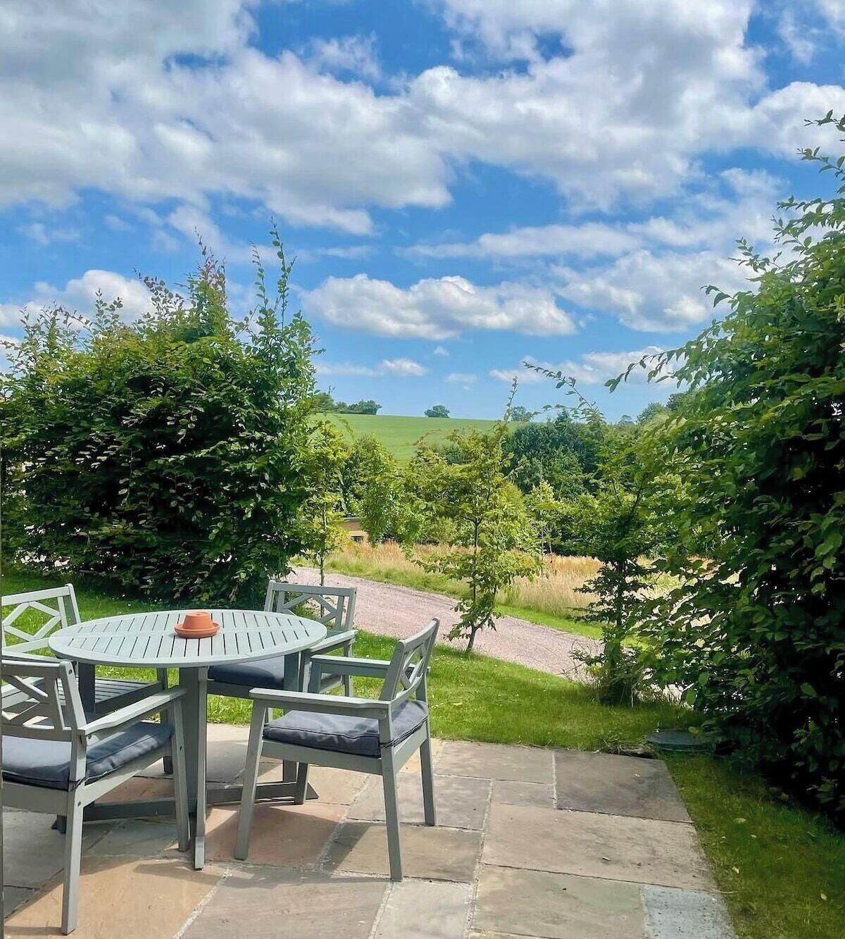 Eine Terrasse mit einem runden Tisch und Stühlen, umgeben von viel Grün und blauem Himmel.