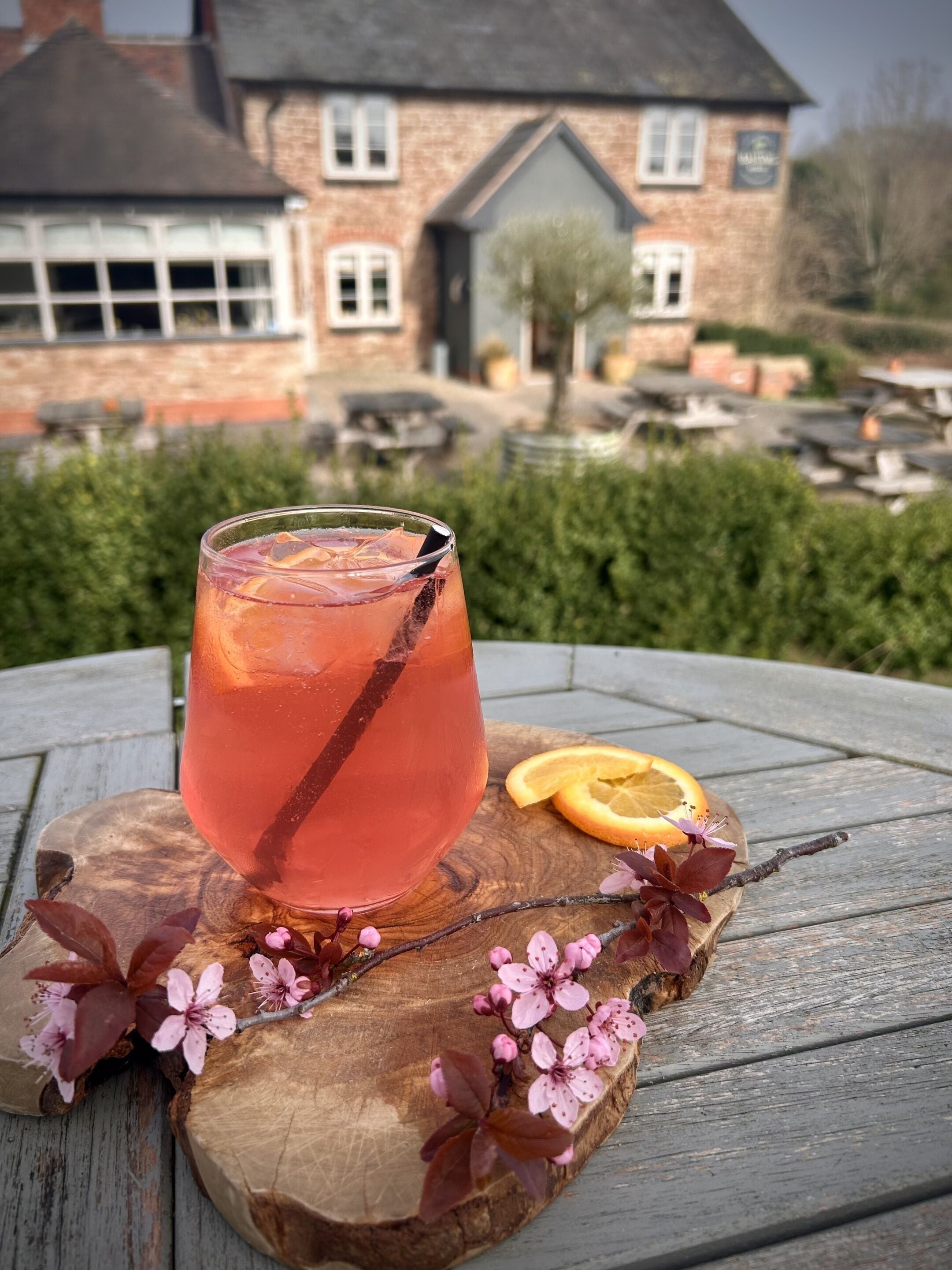 Un vaso de bebida sobre una tabla de madera con flores rosadas, una rodaja de naranja y una casa al fondo.