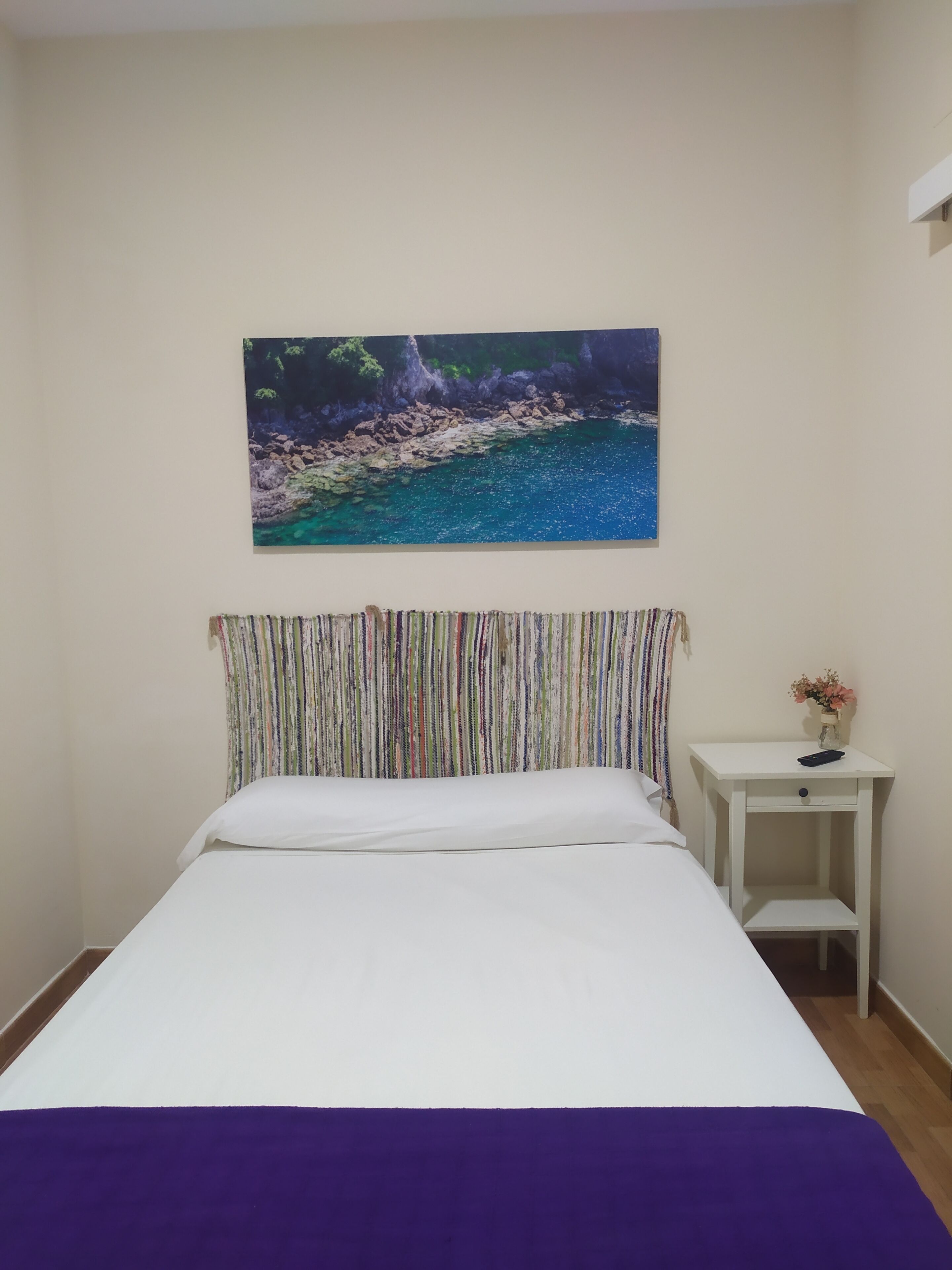 Un dormitorio con cama, mesita de noche, un cuadro y cortina.
