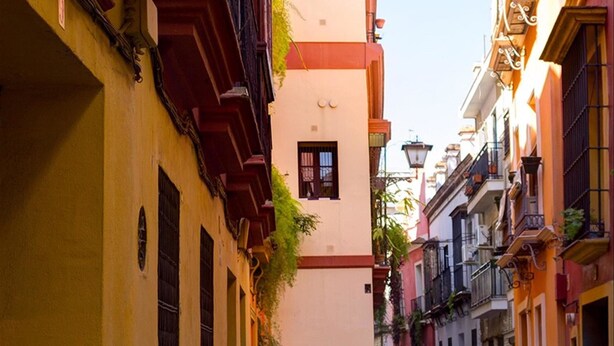 Una calle estrecha flanqueada por edificios, un letrero que dice 'Azahar Pensión Sevilla', y una persona caminando por la calle.