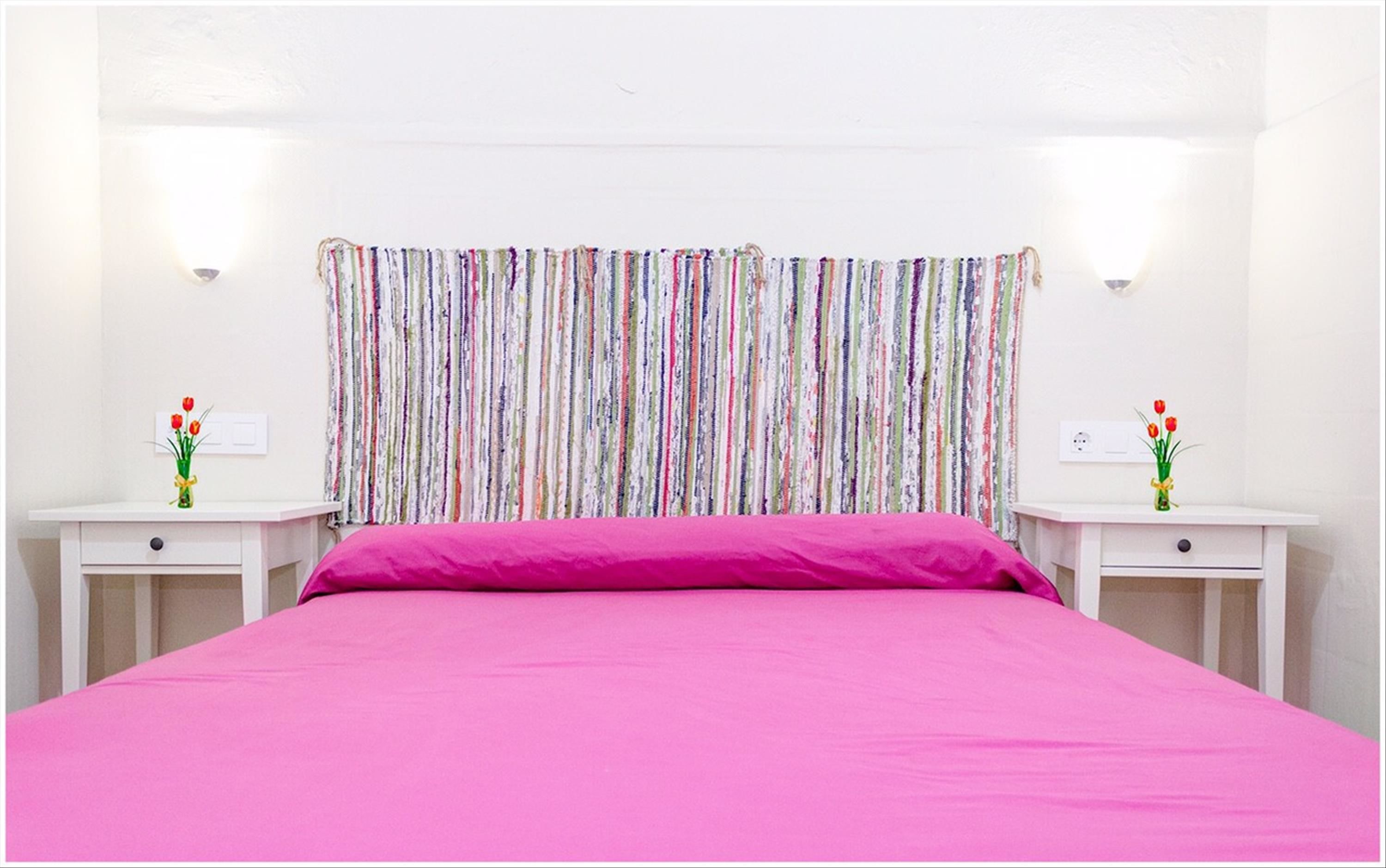 A bedroom with a pink bedspread, striped curtains, and two bedside tables with vases of flowers.