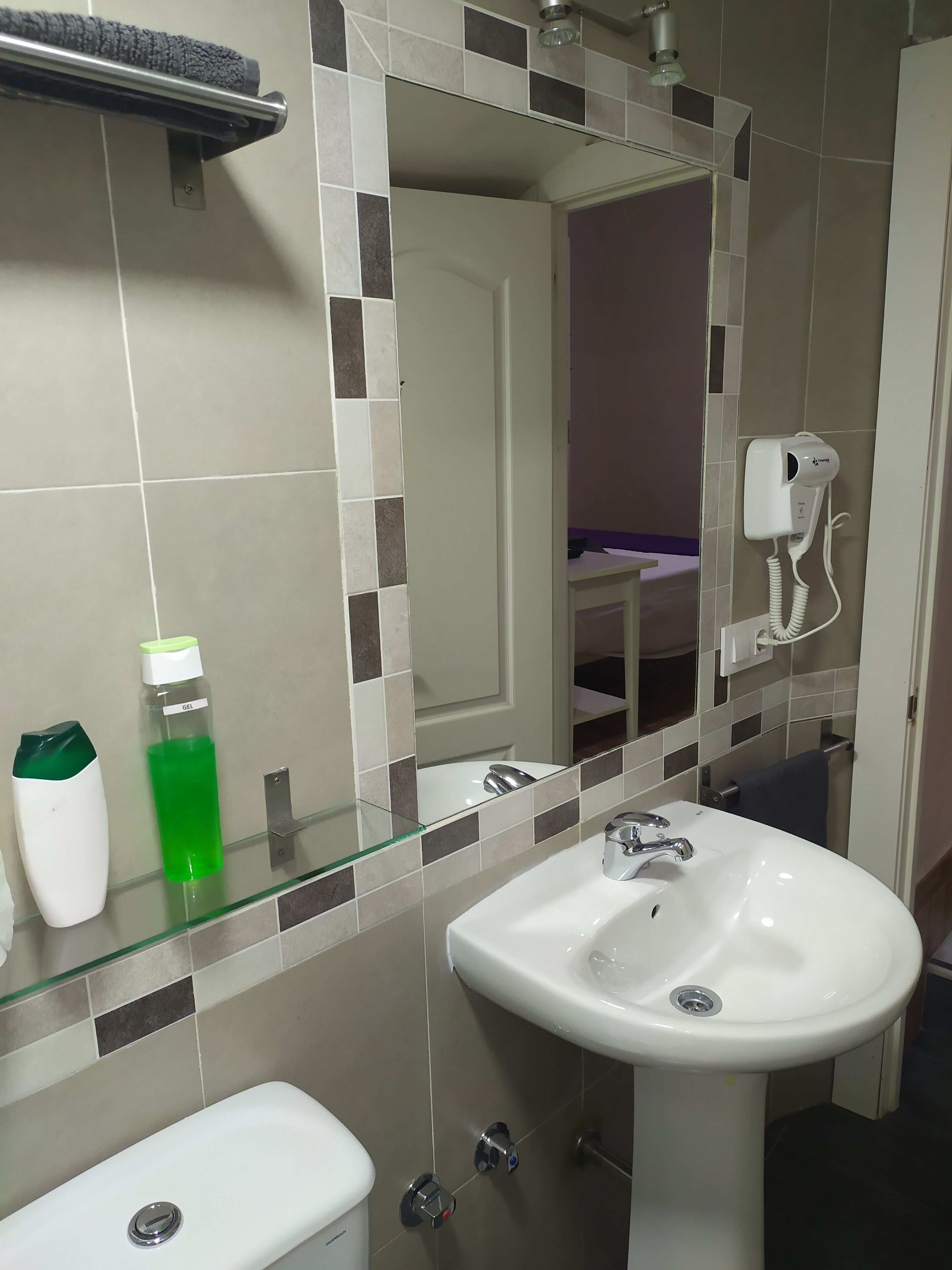 A bathroom with a white sink, a glass shelf with toiletries, a mirror, and a hairdryer on the wall.