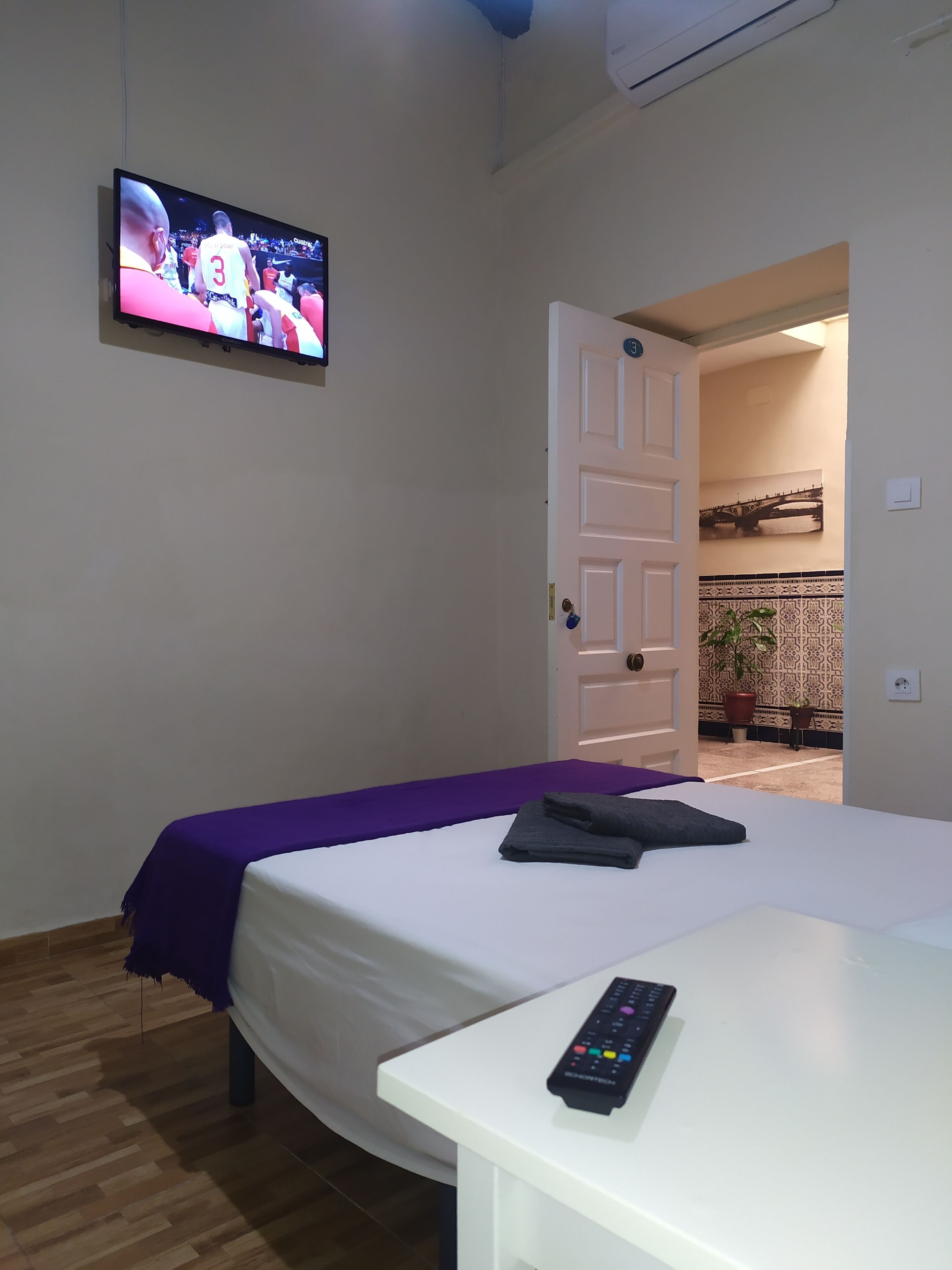 A hotel room with a bed, a TV mounted on the wall, a remote control on the bed, and a door leading to a hallway with a potted plant.