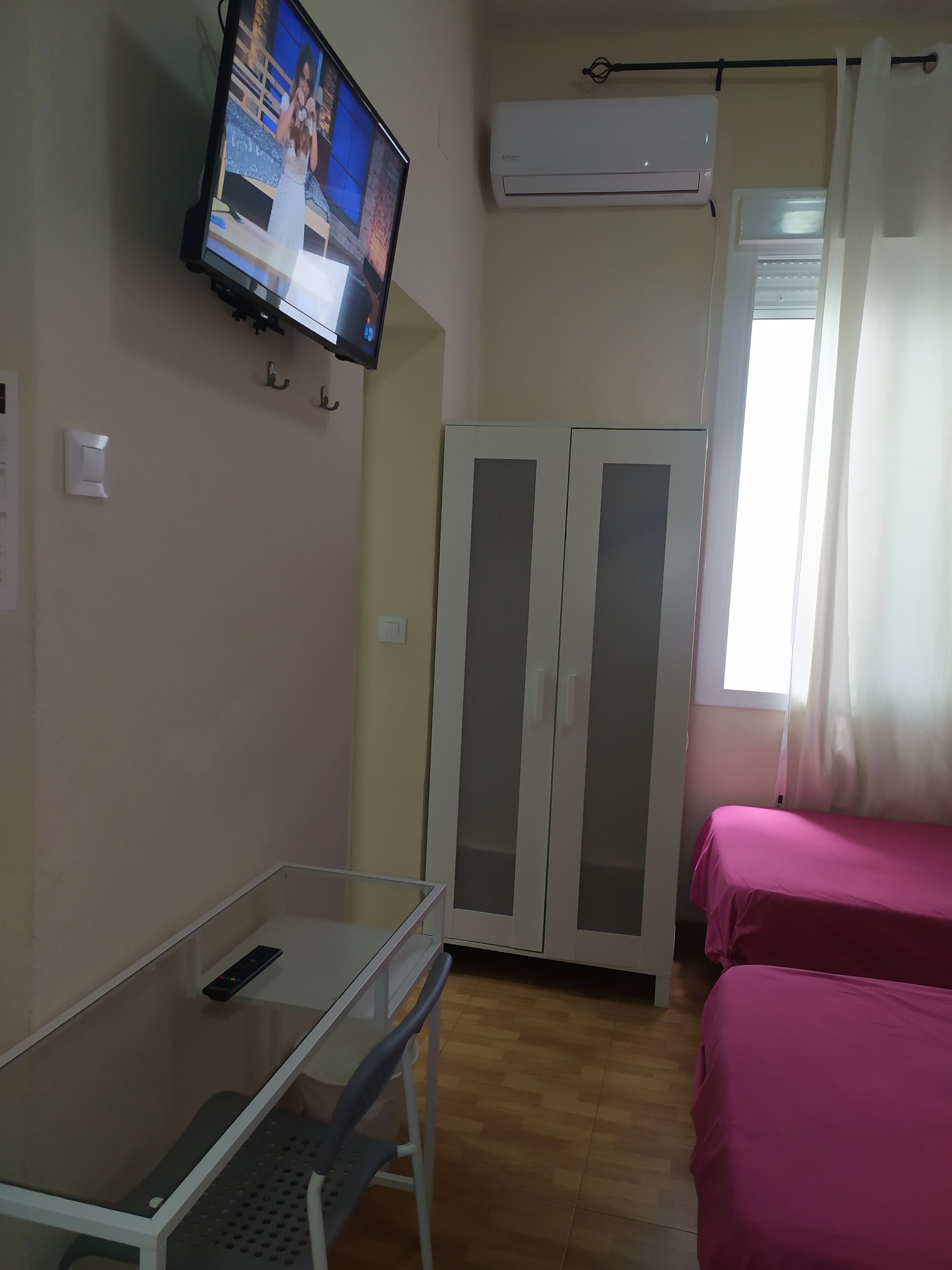 Une chambre avec un téléviseur fixé au mur, une armoire aux portes en verre dépoli, un lit recouvert d’une couvre-lit rose et un appareil de climatisation.