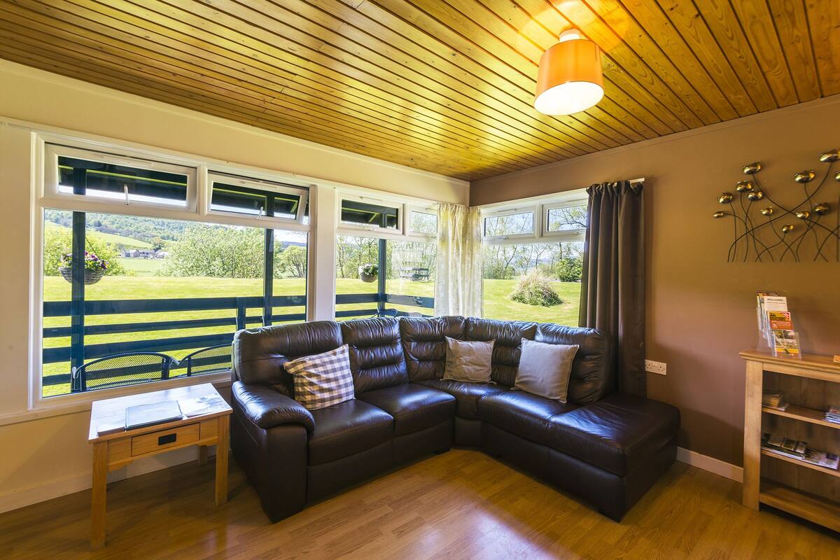 A cozy room with a leather sectional sofa, a small wooden table, and a view of a green landscape through large windows.