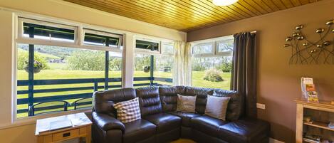 A cozy room with a leather sectional sofa, a small wooden table, and a view of a green landscape through large windows.