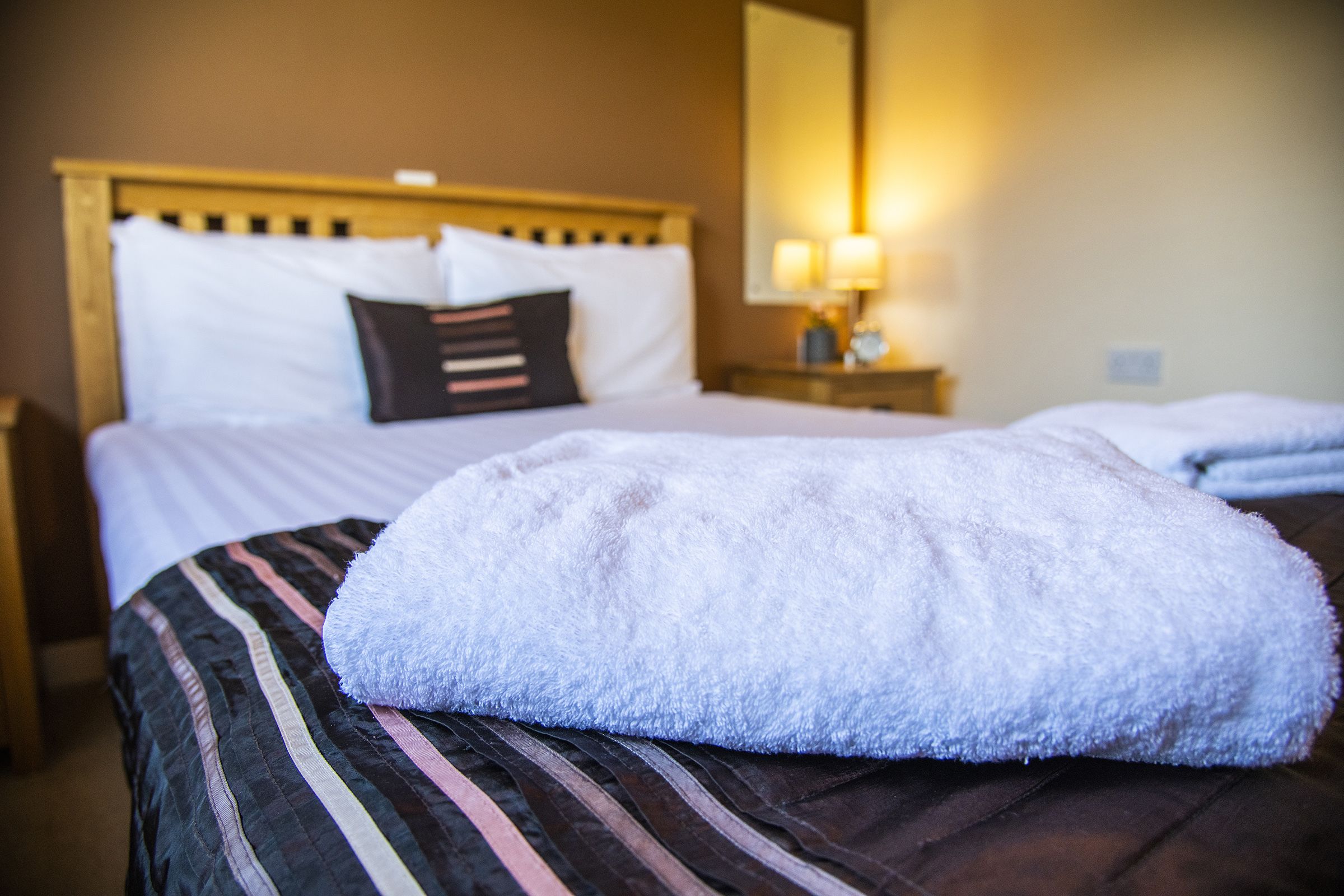 A hotel room with a bed, pillows, a nightstand with a lamp, and a blanket on the bed.
