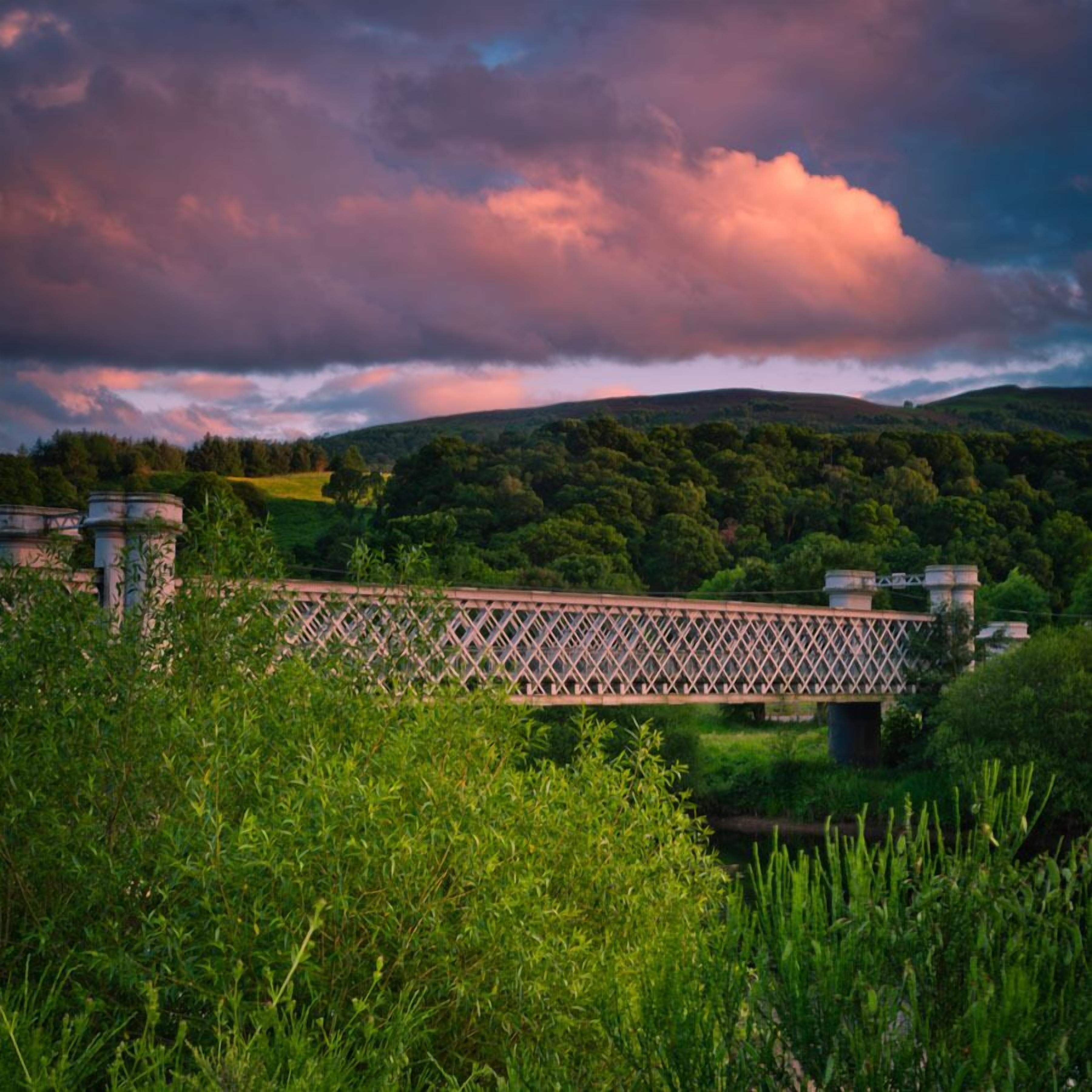 A stone bridge over a river with lush greenery and hills in the background.