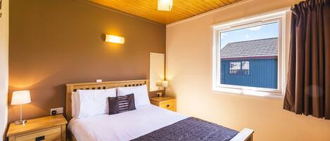 A bedroom with a wooden ceiling, a large bed with white linens, a dark quilt, bedside lamps, and a window with curtains.