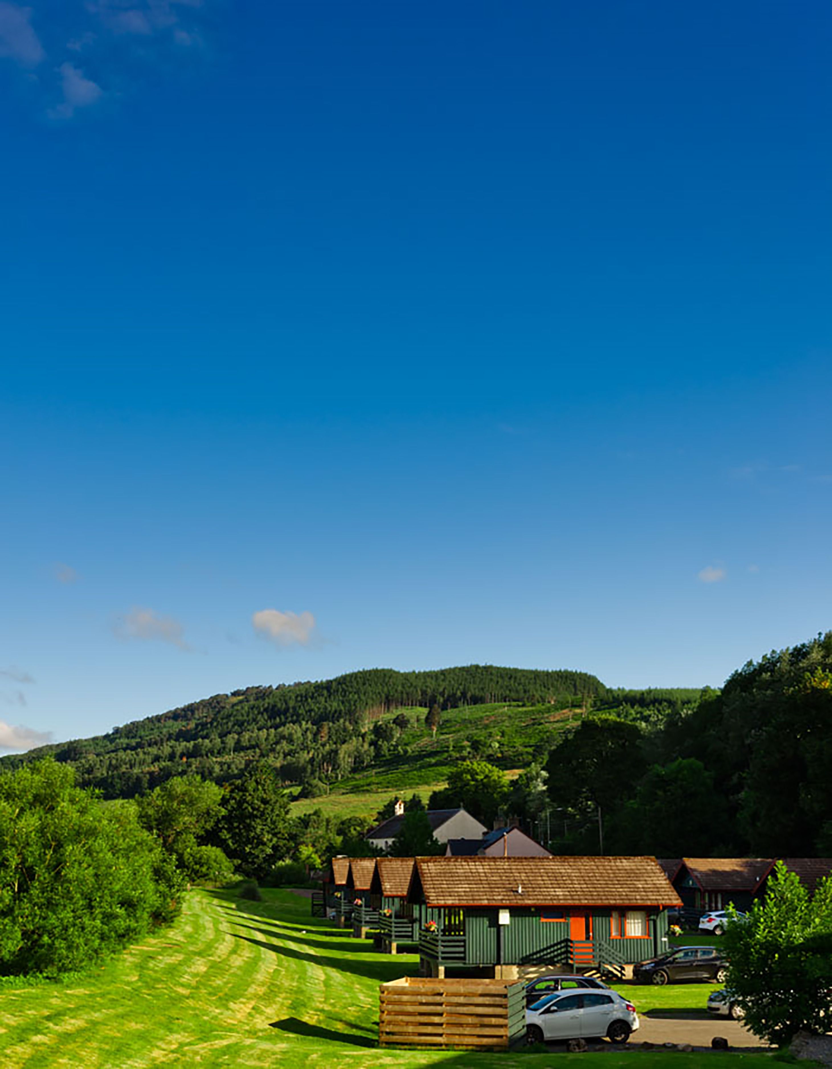 Un paysage rural avec une rangée de bâtiments aux toits verts, un champ d’herbe et une colline en arrière-plan.