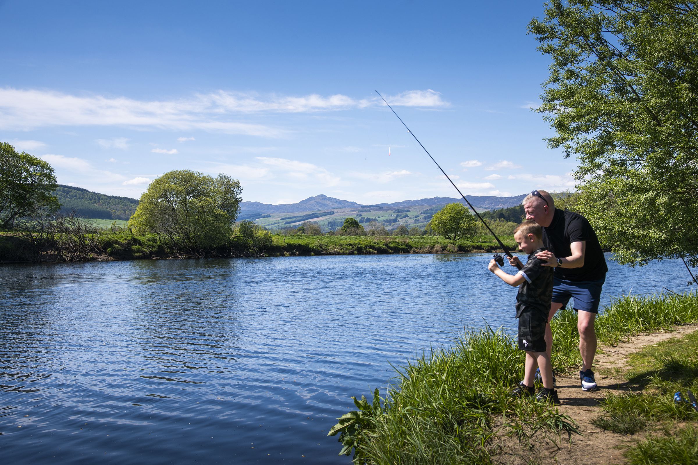 A river with a person fishing, surrounded by greenery and hills.