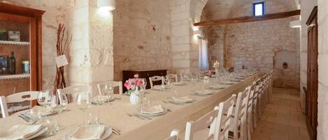 Una larga mesa de comedor dispuesta para una comida en una habitación con paredes de piedra y sillas blancas.