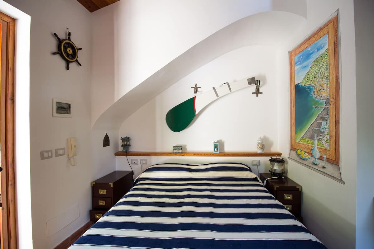 A bedroom with a bed, a wooden headboard, a painting, a wall-mounted light, a telephone, and a small shelf with items.
