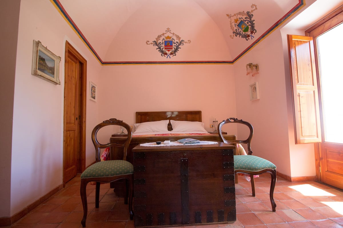A bedroom with a wooden bed, a chest of drawers, two chairs, and a window with sunlight.