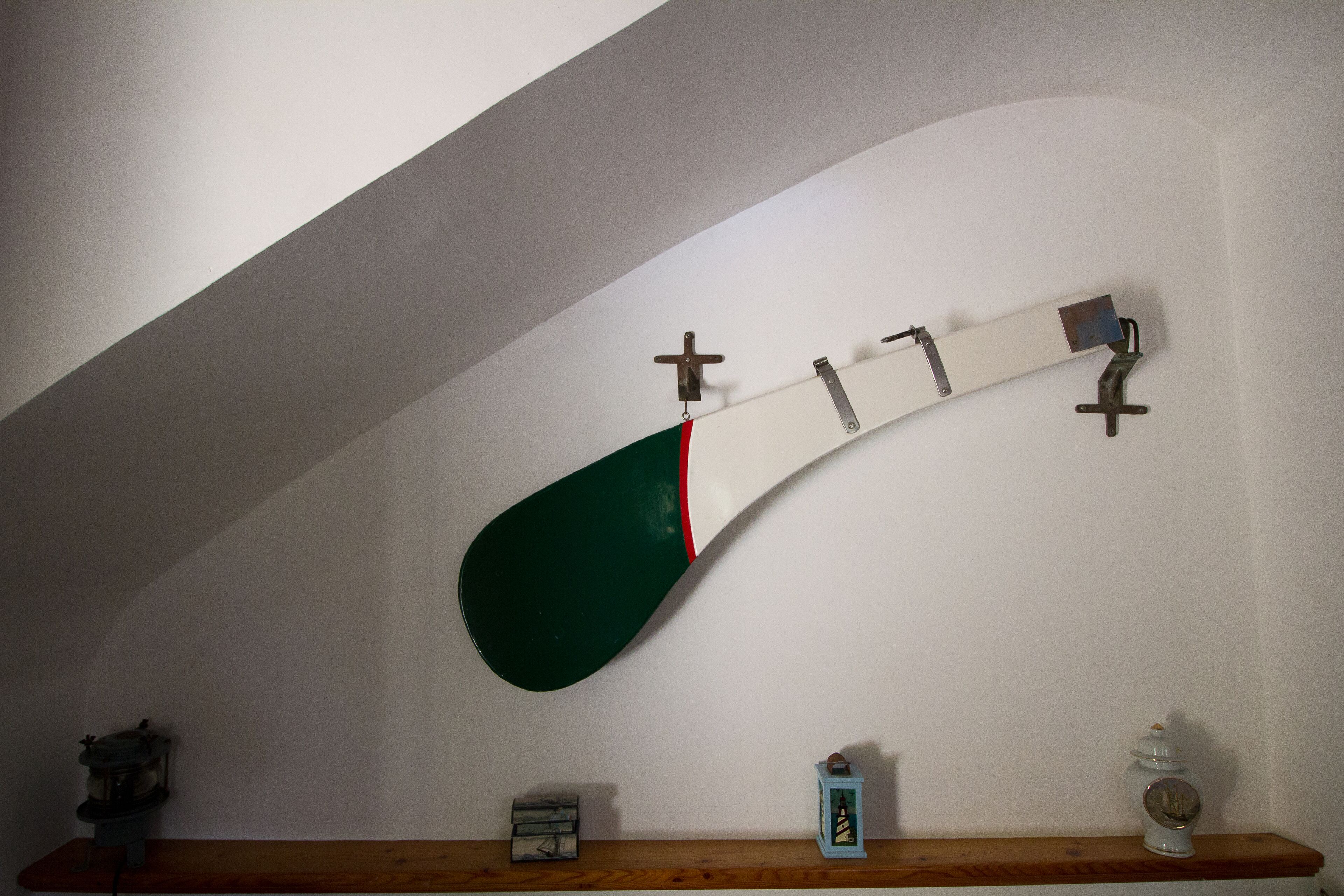 Une planche de skate fixée au mur, avec un motif vert et blanc, une étagère en bois sur laquelle sont disposés divers petits objets, et un mur blanc avec un plafond incurvé.