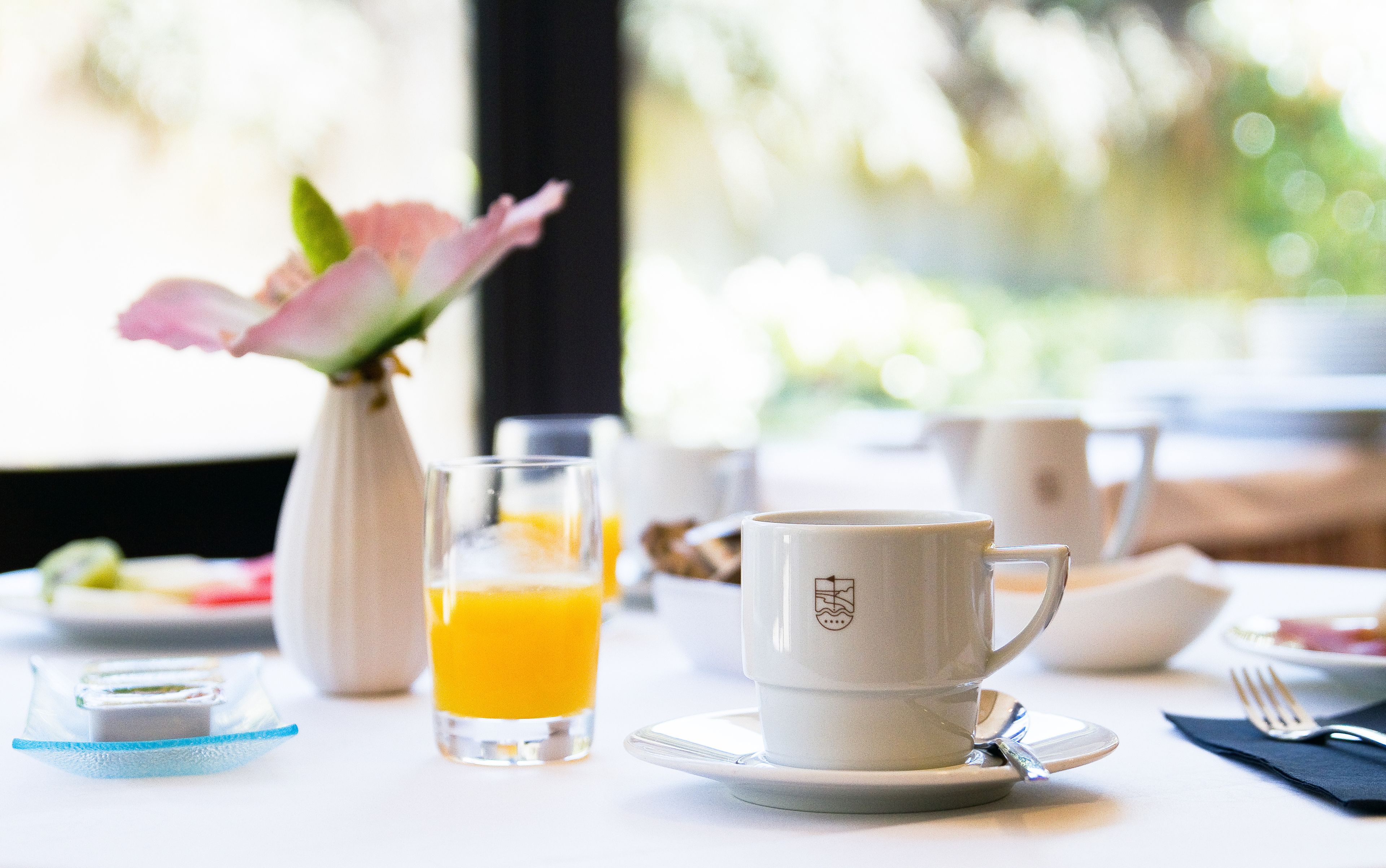 Una mesa de desayuno con una taza, plato, vaso de jugo de naranja y un florero con flores.