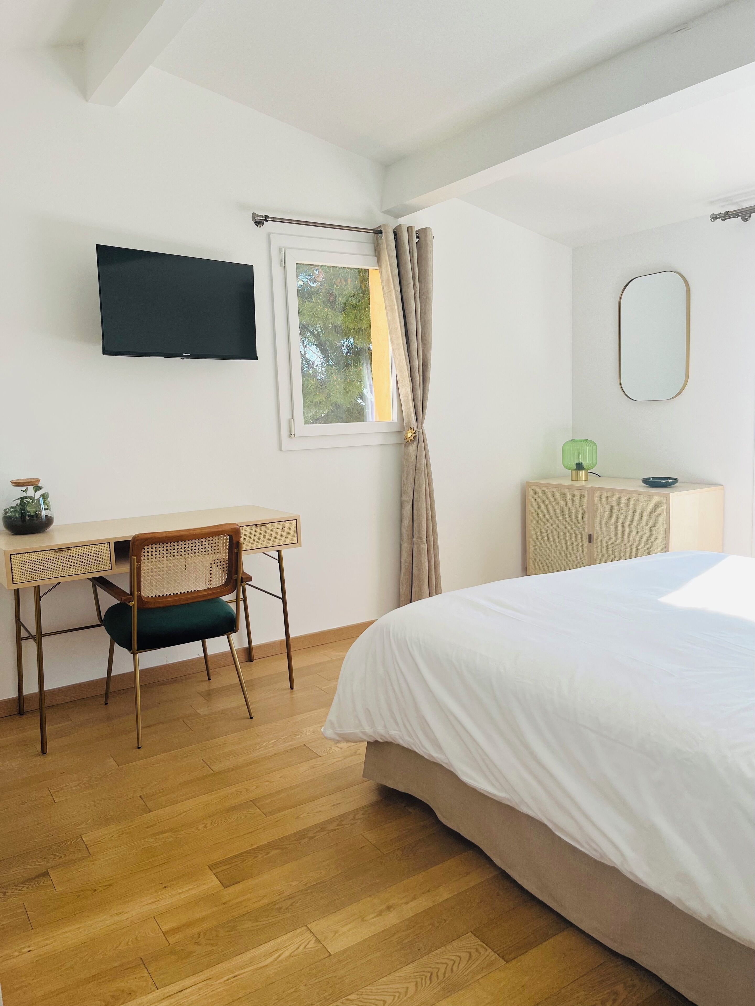 Une chambre à coucher comprenant un lit, un bureau avec une chaise, un téléviseur fixé au mur et une fenêtre avec des rideaux.