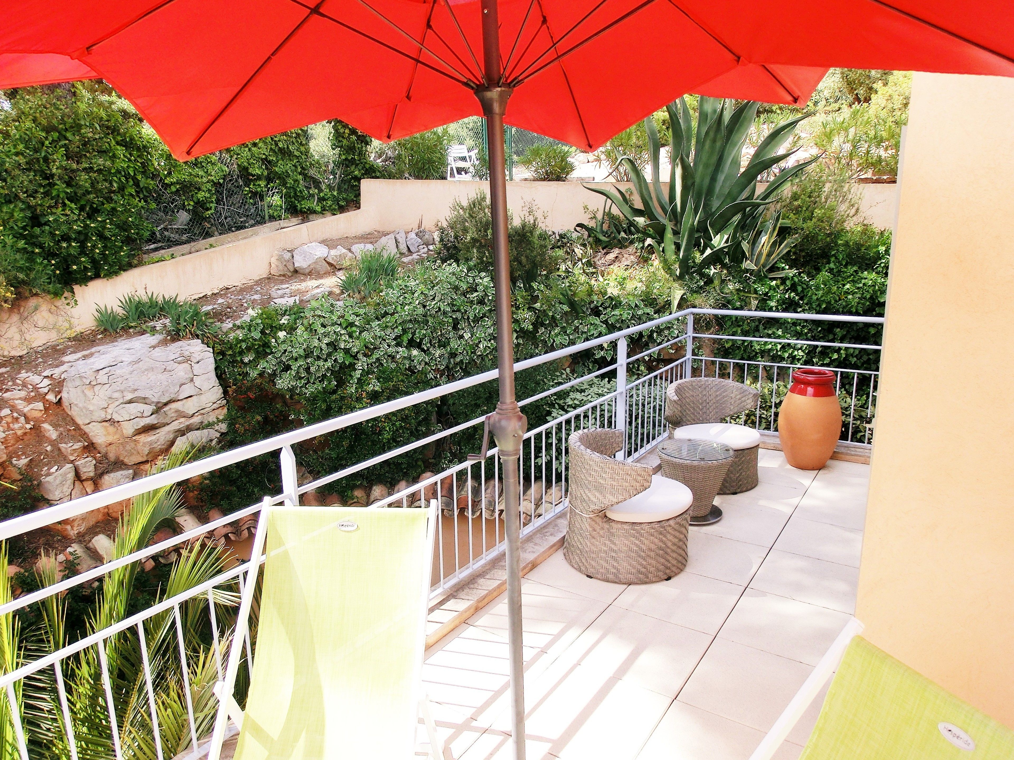 A balcony with wicker furniture, a red umbrella, and a view of a landscaped garden.