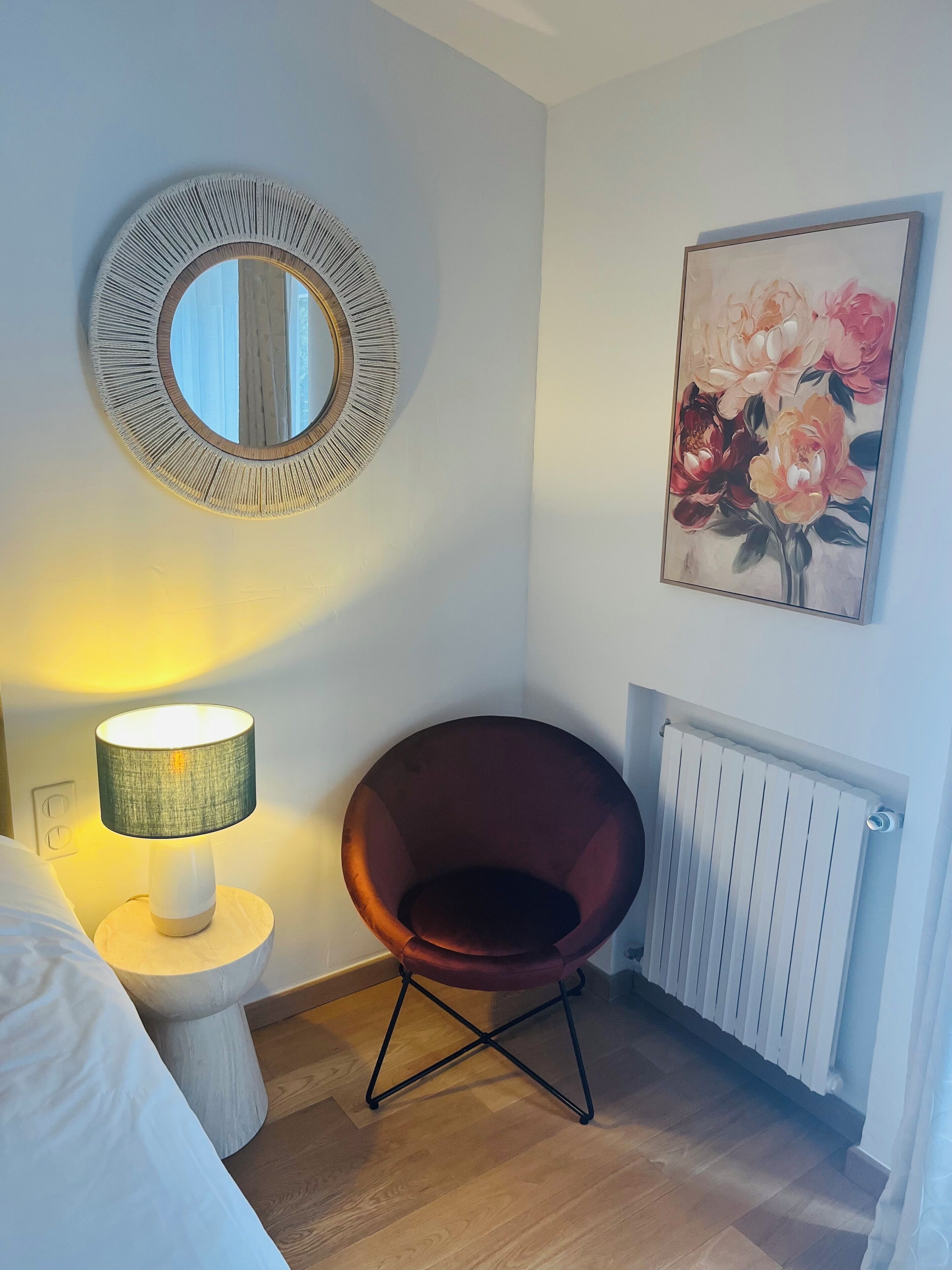 Una habitación con un espejo redondo, un cuadro de flores, una silla roja, una mesita de noche con una lámpara y un radiador.