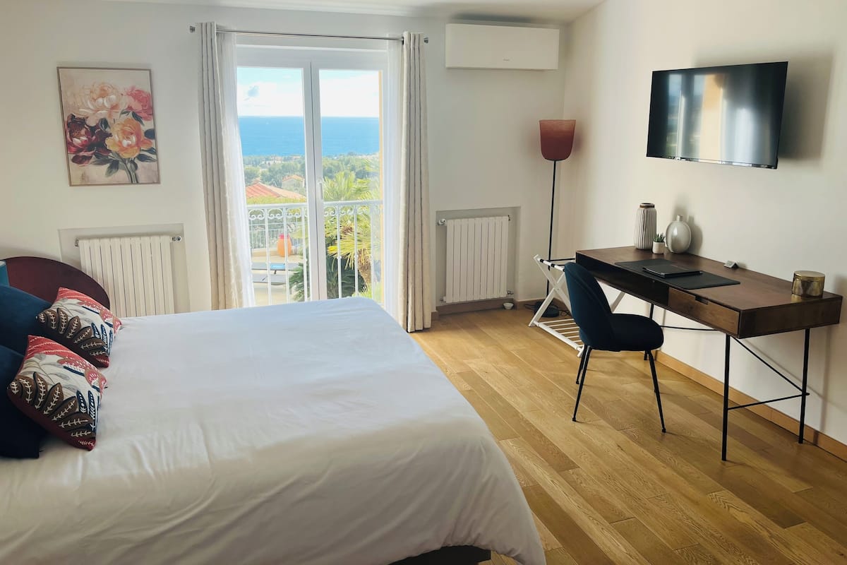 A hotel room with a bed, a desk, a chair, a TV, and a balcony with a view of the sea.