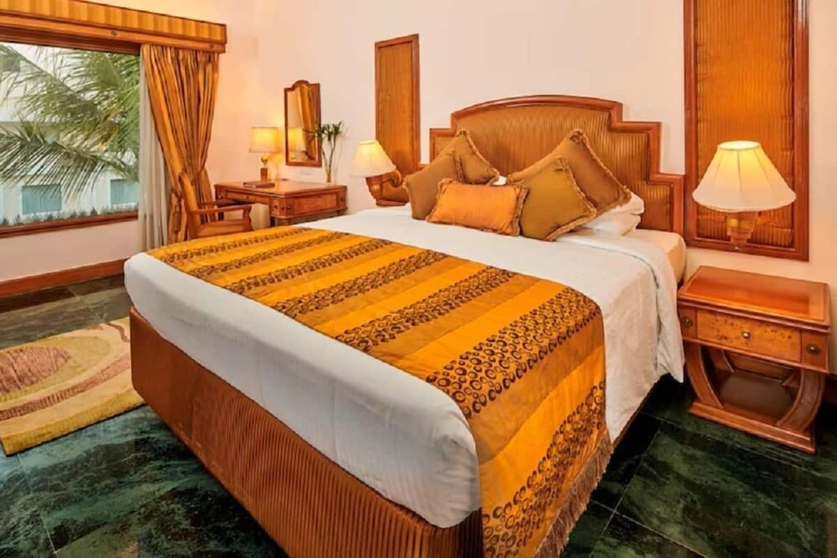 A hotel room with a large bed, wooden headboard, two bedside tables with lamps, a nightstand, a mirror, and a view of palm trees.