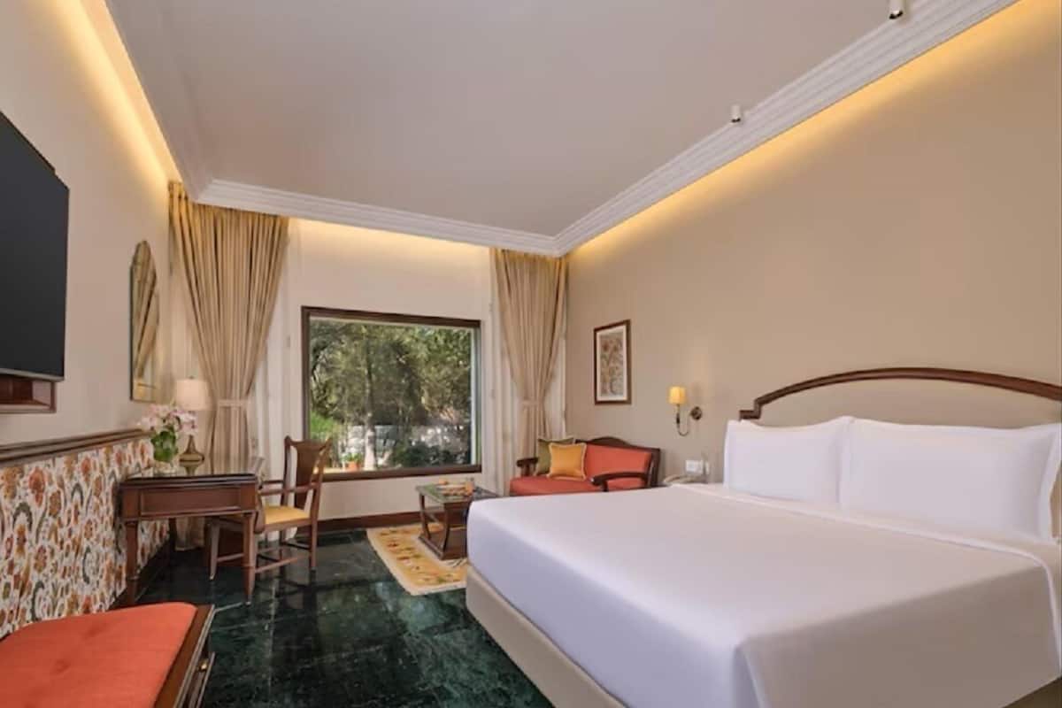 A hotel room with a bed, a desk, a chair, a window with curtains, and a view of greenery outside.