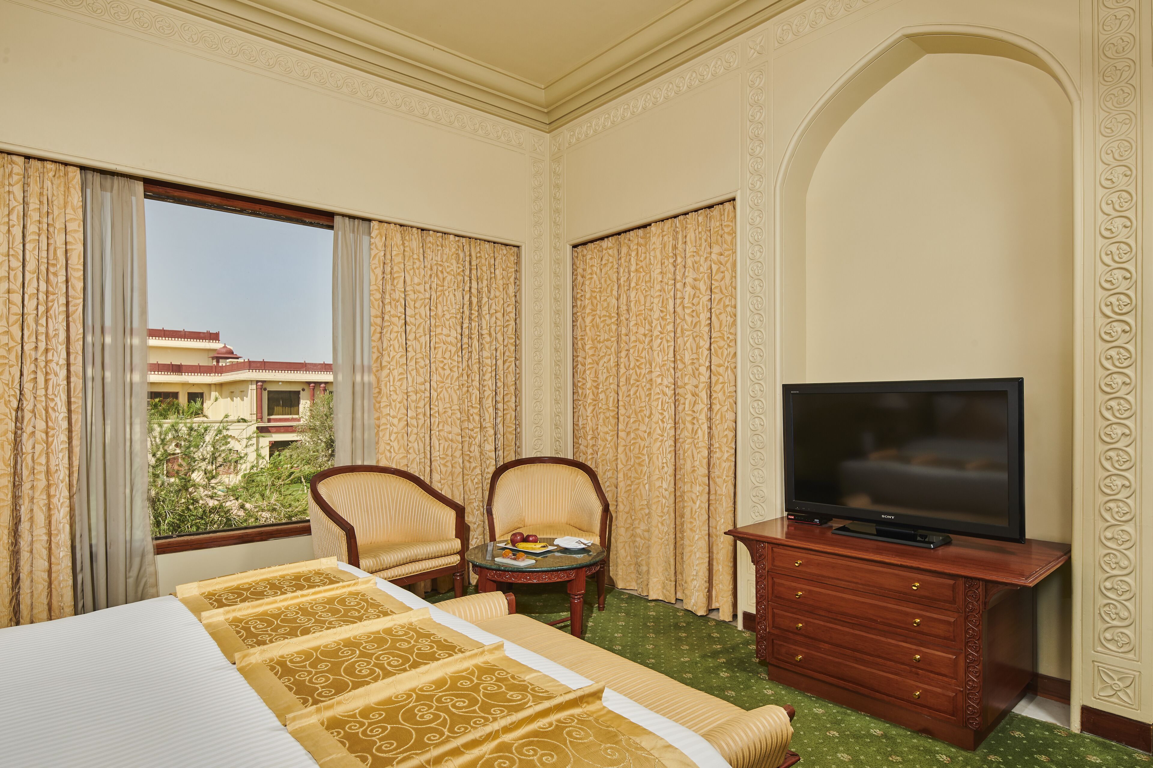 A hotel room with a bed, two chairs, a small table with fruit, a TV on a wooden stand, and a view of a building through the window.