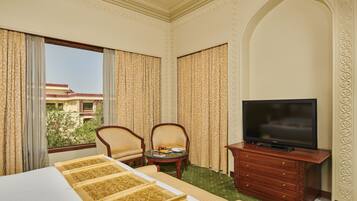 A hotel room with a bed, two chairs, a small table with fruit, a TV on a wooden stand, and a view of a building through the window.