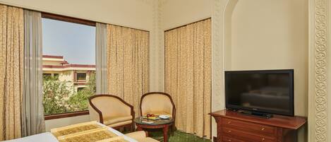 Habitación de hotel con cama, dos sillas, una mesita con fruta, un televisor sobre un mueble de madera y vista a un edificio a través de la ventana.