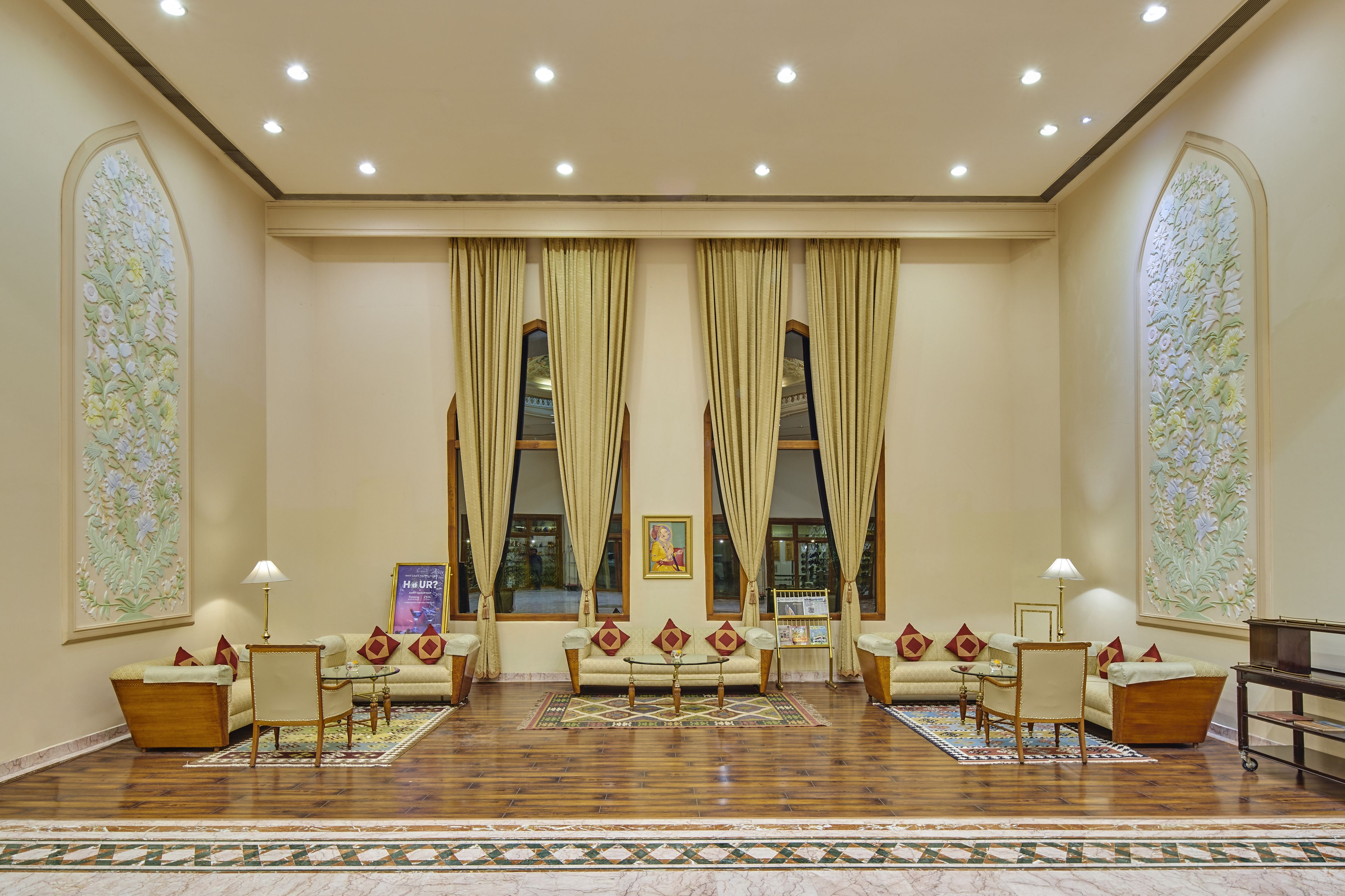 A spacious lobby with a sofa set, a coffee table, and a small table with a lamp. There are two large windows with curtains, a wall-mounted artwork, and a patterned rug on the floor.