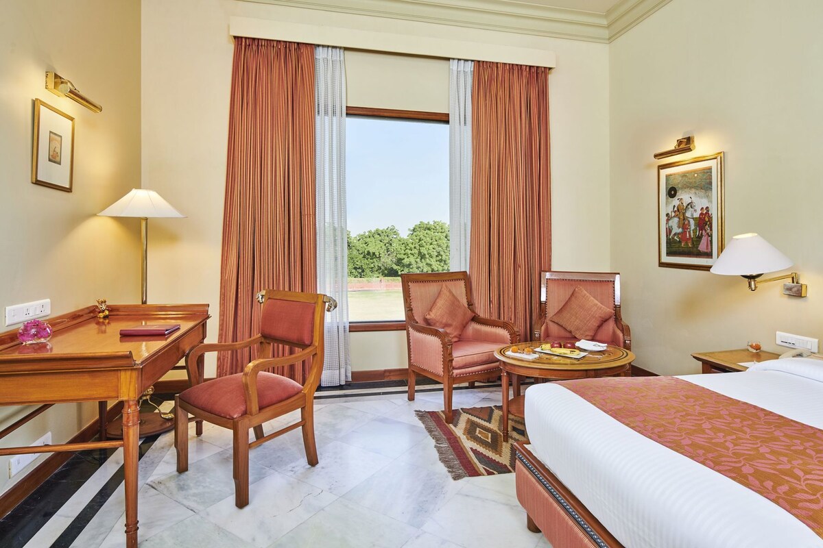 A hotel room with a bed, two armchairs, a desk, a table with food, and a window with curtains.