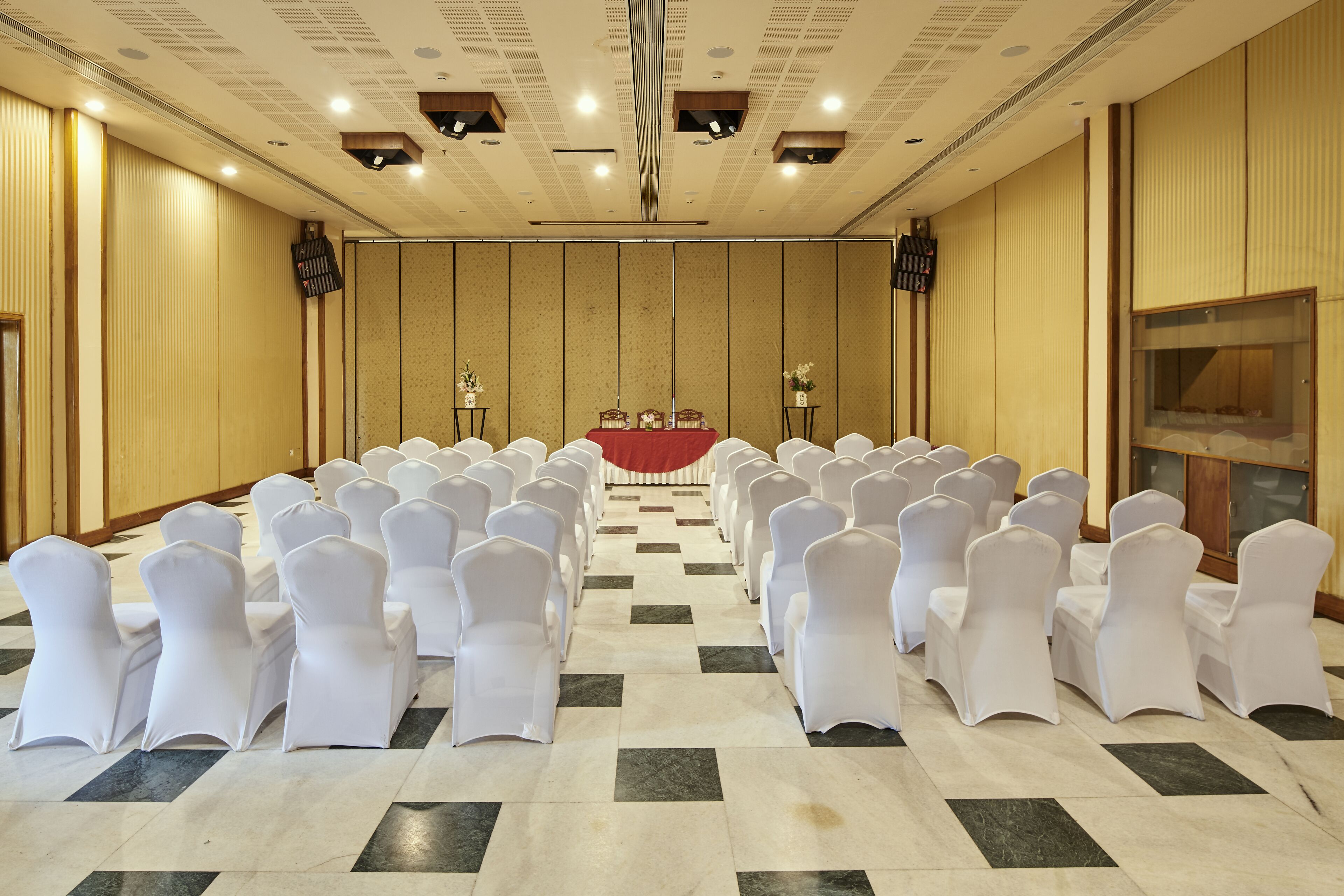 A conference room with rows of white chairs, a red table at the front, and a large screen on the right wall.