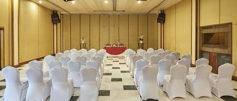 A conference room with rows of white chairs, a red table at the front, and a large screen on the right wall.