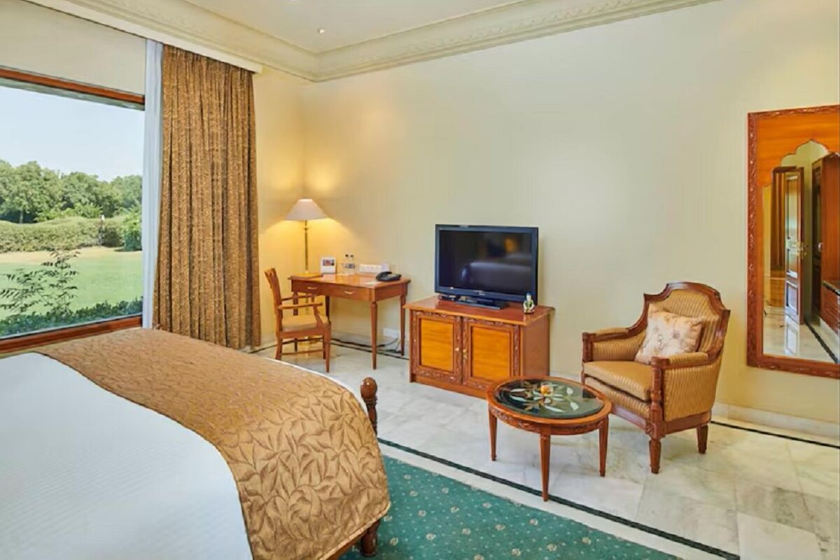 A hotel room with a bed, a desk with a television, a chair, and a view of a green landscape.