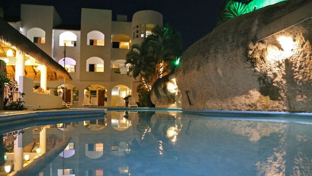 A nighttime view of a hotel pool with clear reflections, palm trees, and a building with arched windows and warm lighting.