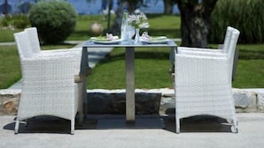 An outdoor dining area with wicker chairs and a table set for two, overlooking a body of water and a grassy landscape. - Belle Helene Hotel (East Mani)