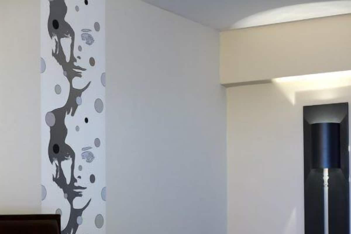Camera d'albergo con un letto, un comodino, una lampada, un cuscino verde e una parete con un motivo astratto grigio.
