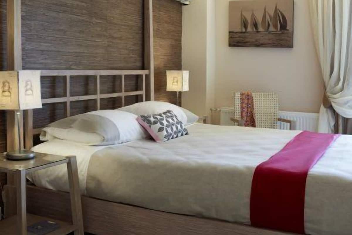 Una camera da letto con testiera in legno, un letto con lenzuola bianche e una coperta rossa, due comodini con lampade, un condizionatore d'aria a parete e un quadro con dei velieri.