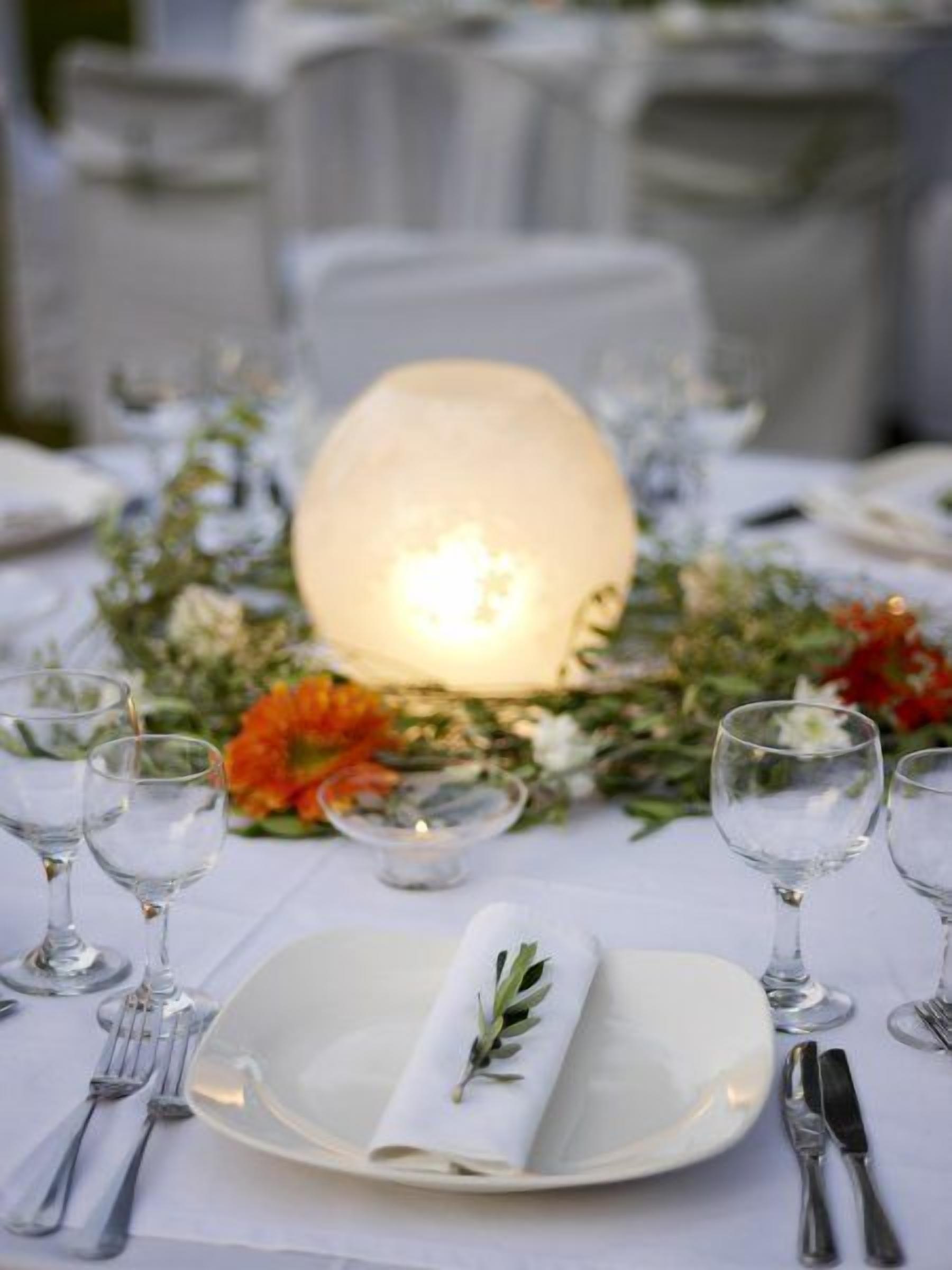Una mesa dispuesta para un evento de cena formal, con un centro de mesa iluminado, copas de cristal y un mantel doblado con un detalle de follaje.
