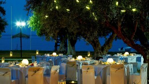 An outdoor dining setup with tables set for a meal, surrounded by trees and a view of the sea. - Belle Helene Hotel (East Mani)