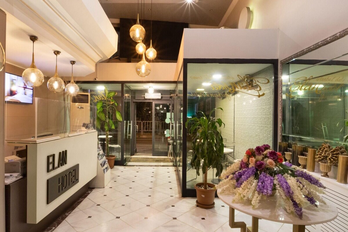 A hotel reception area with a reception desk, a large potted plant, and a table with a floral arrangement.